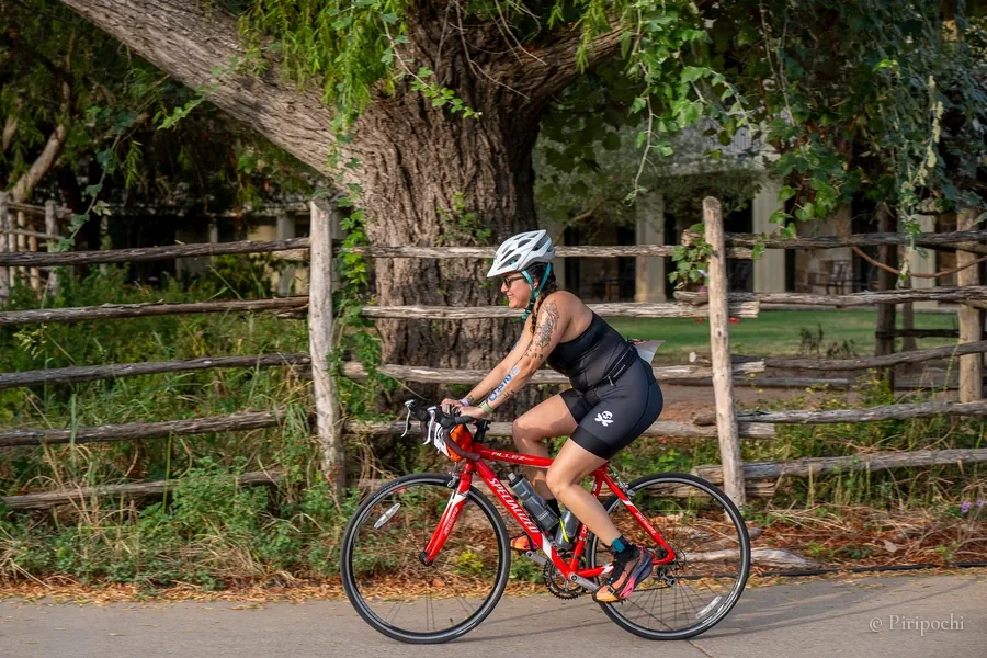Atleta de triatlón pedaleando una bicicleta de ruta roja junto a una cerca de madera y un gran árbol