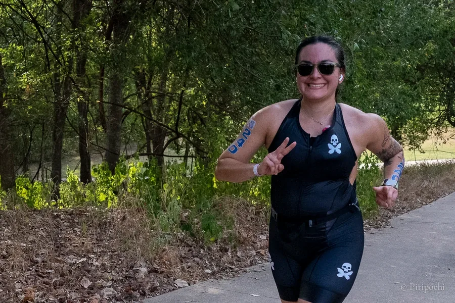 Atleta de triatlón corriendo sonriente y haciendo la señal de paz en un sendero entre árboles